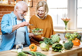 An older couple standing in their kitchen and using leafy greens and vegetables to make a healthy salad